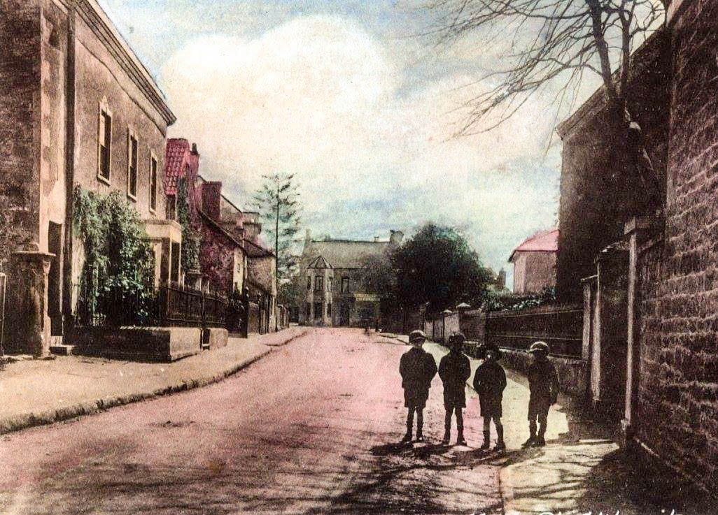 A hand-coloured view with four children standing in the middle of the road. The colour is an early colourist's guess, not a record.