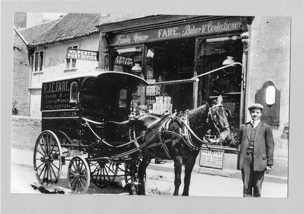 The shopfront with its window display, a horse-drawn delivery van at the kerb and a man standing at the horse's head.