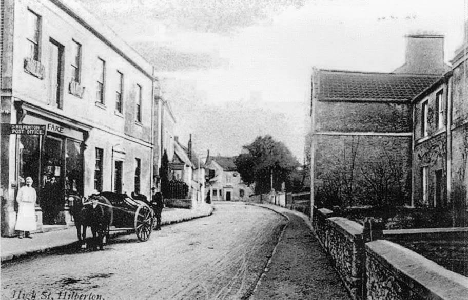 A horse and cart standing outside a village shop, with a figure in a long apron in the doorway.