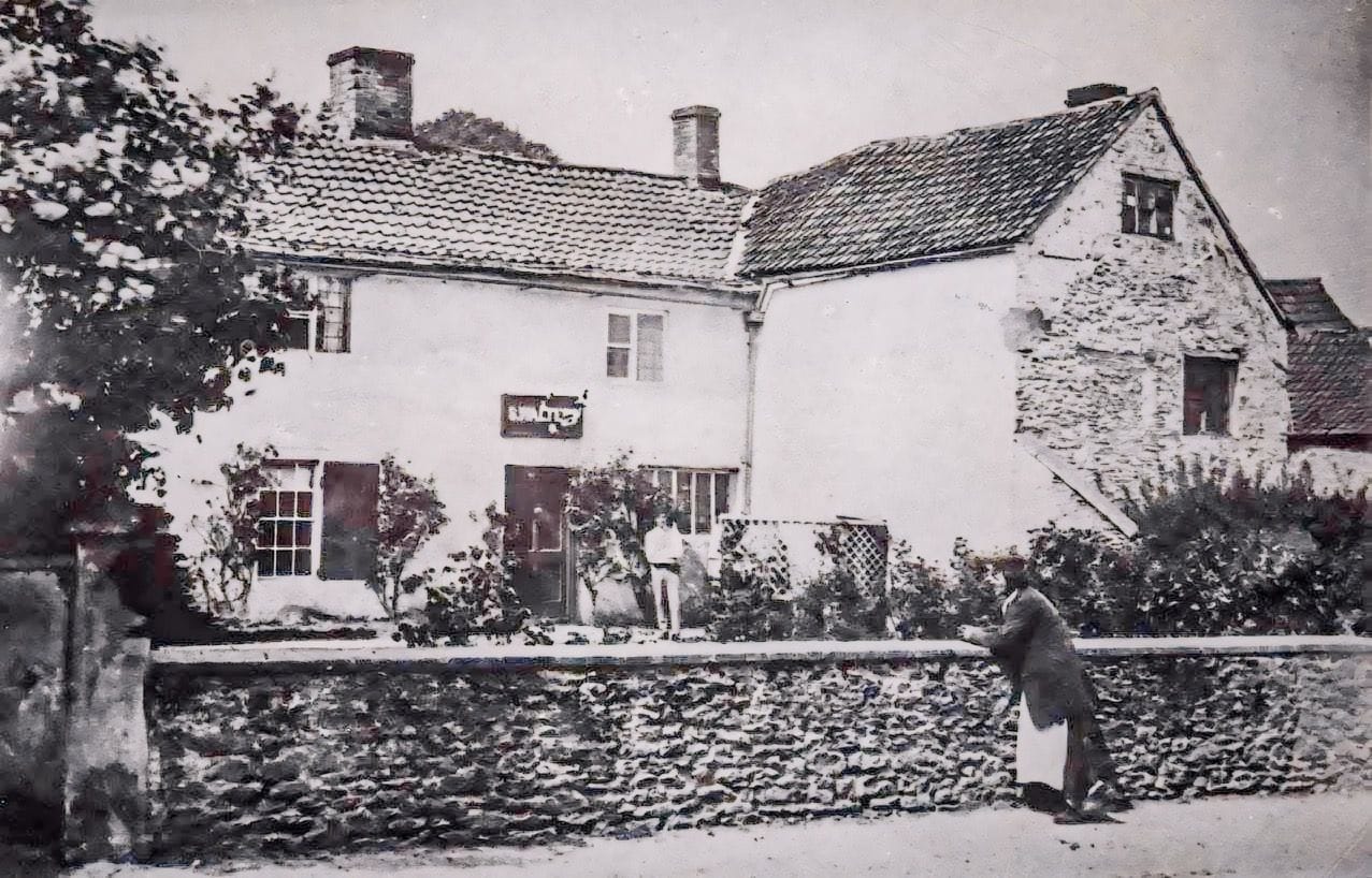 The surviving photograph on which the reconstruction is based: a rendered range with a tiled roof, an adjoining stone gable, a sign above the door and a rubble boundary wall.