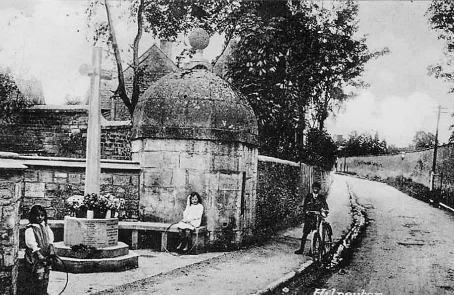 A stone monument set against a wall, with a lane running away beside it and two figures in the distance.